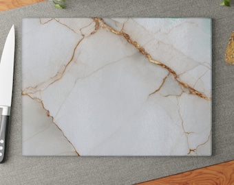 Puede incluir: Una tabla de cortar rectangular con un patrón de mármol blanco y vetas doradas prominentes que recorren la superficie en diagonal. La tabla tiene un acabado liso y pulido, y está colocada junto a un cuchillo de cocina plateado.