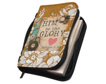 Puede incluir: Una funda para libro con cremallera en color amarillo mostaza con ilustraciones florales blancas y un gráfico central con el texto "to HIM be the GLORY" y "ROMANS 11:36" junto a un pequeño corazón rojo, un reloj vintage y detalles en azul cerceta.