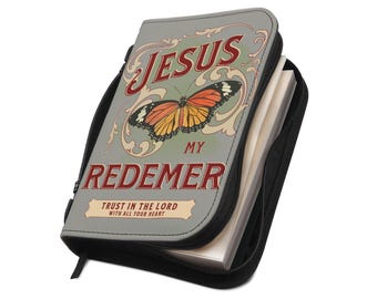 Puede incluir: Una funda gris para Biblia con cremallera que presenta una ilustración de mariposa monarca y el texto "JESUS MY REDEMER" en letras rojas, con una banda en la parte inferior que muestra el texto "TRUST IN THE LORD WITH ALL YOUR HEART" en negro.