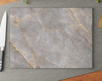 Puede incluir: Una tabla de cortar rectangular con un patrón de mármol gris claro y vetas doradas finas e intrincadas por toda la superficie. La tabla tiene un acabado liso y pulido y está colocada junto a un cuchillo de cocina con mango negro.