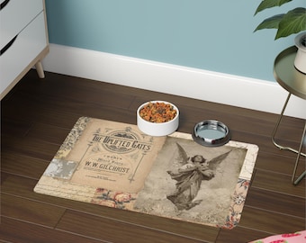 May include: A beige pet feeding mat with a vintage design featuring an angel with wings and the text "The Uplifted Gates" with a floral border. The mat is on a wooden floor with a white bowl of pet food and a silver water bowl.