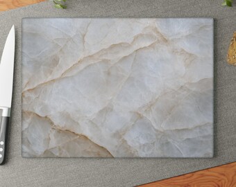 Puede incluir: Una tabla de cortar rectangular que presenta un patrón de mármol blanco y gris claro con vetas de color tostado sutiles en toda la superficie. La tabla tiene un acabado liso y pulido y está colocada horizontalmente con un cuchillo a la izquierda.