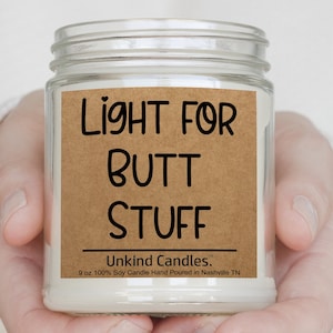 May include: A clear glass jar candle with a tan paper label featuring black text that reads LIGHT FOR BUTT STUFF, Unkind Candles, and 9 oz 100% Soy Candle Hand Poured in Nashville TN. The candle contains white wax and has a visible wick at the top.