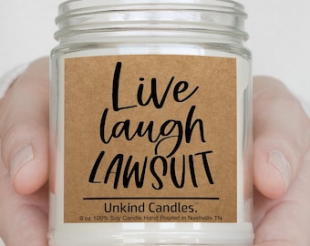May include: A clear glass jar candle filled with white wax. The label is brown paper with black text that reads: Live laugh LAWSUIT, Unkind Candles, 9 oz 100% Soy Candle Hand Poured in Nashville TN. The jar has a threaded top and a smooth finish.