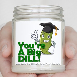 Peut inclure: Une bougie en pot de verre transparent avec une étiquette blanche représentant un cornichon de dessin animé portant une toque de diplômé et faisant un pouce levé. Le texte "You're A Big Dill!" est en vert. La bougie est de 227 grammes.
