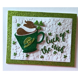 May include: A St. Patrick's Day greeting card with a green glitter border. The card features a green mug with a leprechaun hat, a shamrock, and the phrase "luck of the irish" in green script. The background has a white clover pattern.