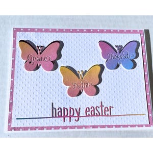 May include: A greeting card with three butterfly cutouts in shades of pink, yellow, and blue, each with a word: "Grace", "Faith", and "Blessed". The words "happy easter" are cut out at the bottom, with a pink dotted border.