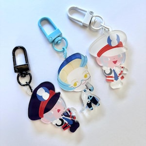 Op de afbeelding: Drie acryl sleutelhangers met cartoonkarakterontwerpen. Eén sleutelhanger heeft een personage met een marineblauwe hoed en uniform, een andere heeft blauw haar en een bril, en de derde heeft een witte hoed en uniform. Elke sleutelhanger heeft een verschillend gekleurde sluiting.