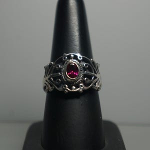 May include: A silver ring with an oval, red gemstone at the center. The ring has an ornate, filigree design around the gemstone and band. The ring is displayed on a black ring holder against a neutral background.