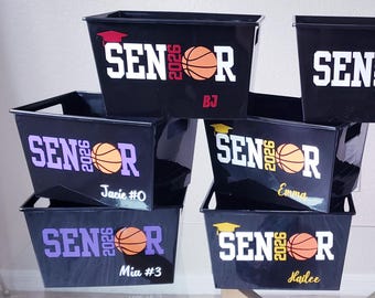 Puede incluir: Siete cajas de almacenamiento rectangulares de plástico negro con el texto SENIOR 2026 y un gráfico de baloncesto que sustituye a la letra O. Cada caja está personalizada con un nombre único en varios colores como blanco, morado y amarillo.
