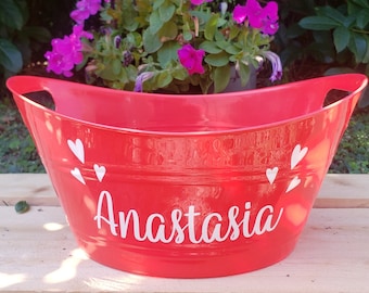 Op de afbeelding: Een rode metalen bak met gebogen zijkanten en handvatten. Witte harten en de naam "Anastasia" staan op de voorkant. De achtergrond toont een houten oppervlak en paarse bloemen.