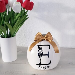 May include: A white ceramic pumpkin featuring a black letter E with a leaf design and the name Enzo printed below it. The pumpkin is topped with a shiny gold ribbon bow. Beside it stands a white vase filled with red and white tulips.