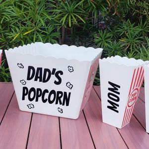 May include: A set of white popcorn containers with red stripes and black text. The large container reads "DAD'S POPCORN". The smaller containers read "ALEXIS", "MOM", and "BRADY". The containers are on a pink surface.