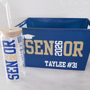 May include: A clear glass tumbler with a bamboo lid and straw, and a navy blue bin. Both items are decorated with the word "SENIOR" and the year "2026" in gold and navy blue. The bin also has the name "Taylee #31".