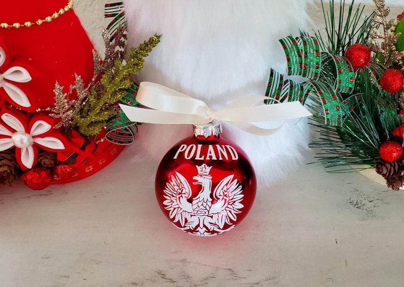 Op de afbeelding: Een rode kerstbal met het woord "POLAND" in witte letters. De ornament heeft een wit Pools adelaarsymbool. Het is vastgebonden met een cr&egrave;mekleurig lint. Feestelijke vakantiedecoratie op de achtergrond.