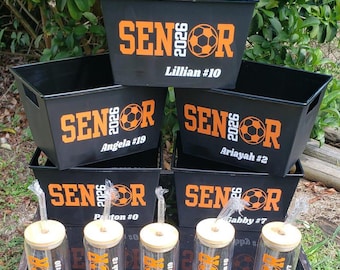May include: Five black plastic storage bins and five clear glass tumblers with bamboo lids and straws. Each item features orange text reading SENIOR 2026 with a soccer ball graphic and individual names like Lillian #10, Angela #19, and Ariayah #2.