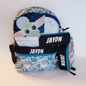 Pode incluir: Um conjunto de mochila, estojo e lancheira com tema Baby Yoda. A mochila e a lancheira apresentam um padr&atilde;o azul e turquesa com Baby Yoda e outros personagens. O nome "JAYDN" est&aacute; escrito em branco no estojo e na mochila.