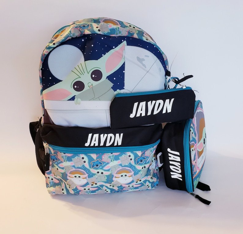 Pode incluir: Um conjunto de mochila, estojo e lancheira com tema Baby Yoda. A mochila e a lancheira apresentam um padr&atilde;o azul e turquesa com Baby Yoda e outros personagens. O nome "JAYDN" est&aacute; escrito em branco no estojo e na mochila.