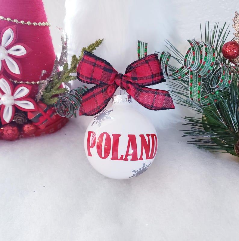Op de afbeelding: Een witte kerstbal met het woord "POLEN" in rode letters. De ornament is versierd met zilveren sneeuwvlokken en een rood-zwarte ruitstrik. Feestelijke vakantiedecoratie.