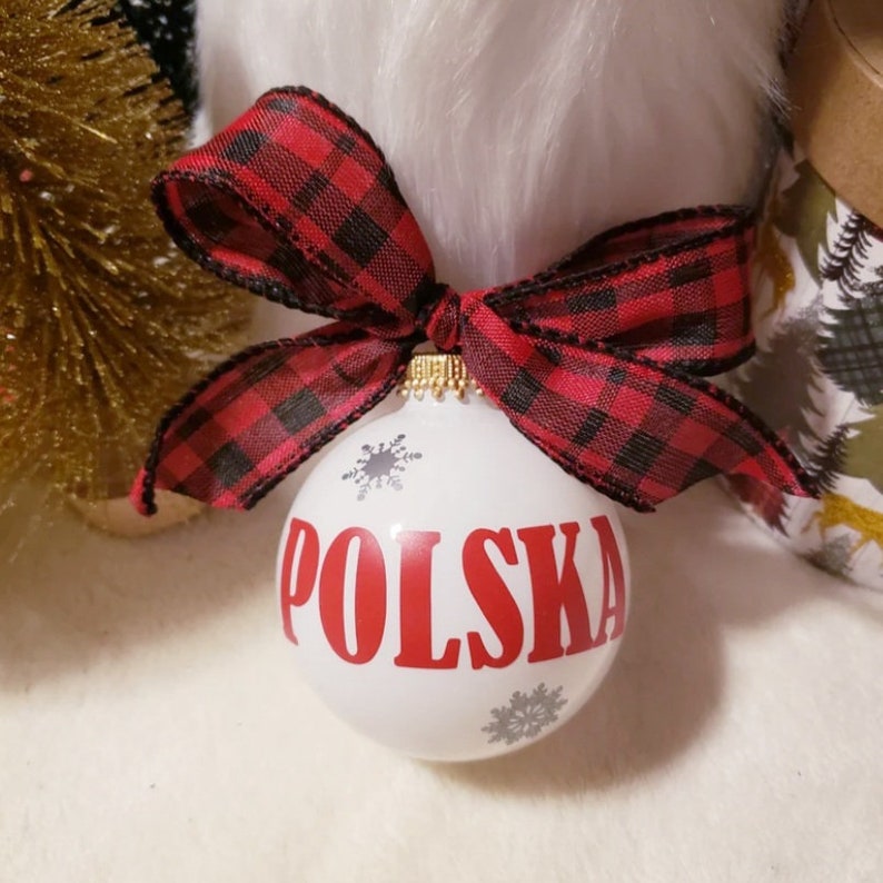 Op de afbeelding: Een witte kerstbal met het woord "POLSKA" in rode letters en grijze sneeuwvlokaccenten. De bal is versierd met een rood-zwarte geruite strik. Een feestelijke vakantieversiering.