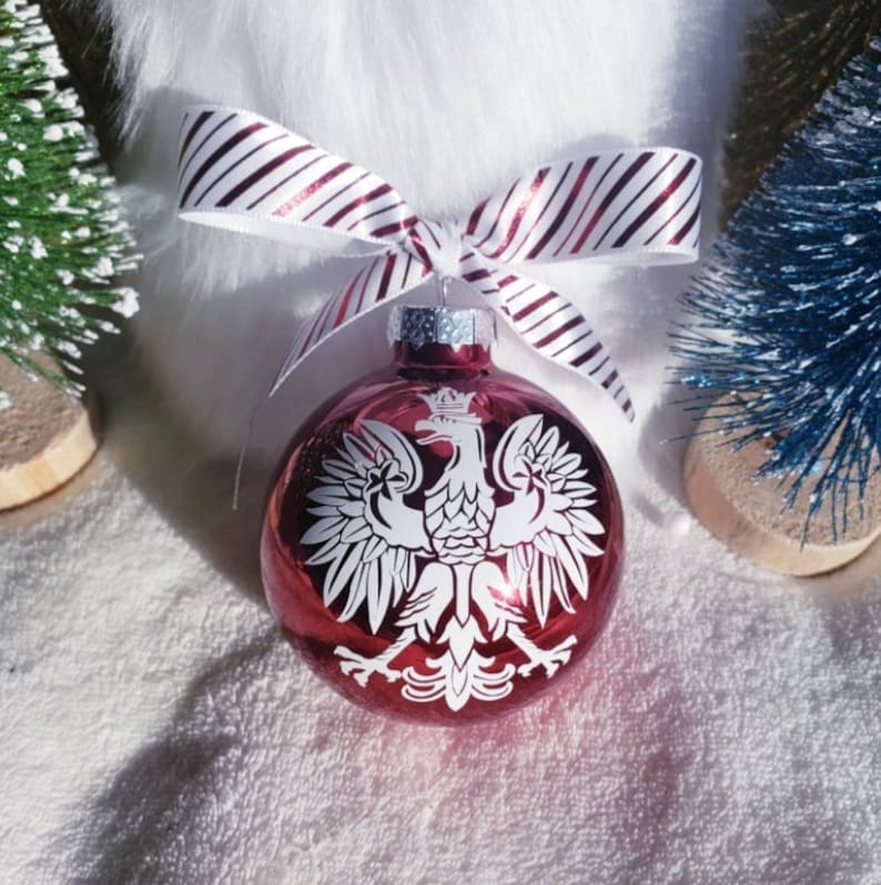 Op de afbeelding: Rode kerstbal met een wit Pools adelembleem. De bal heeft een zilveren dop en is versierd met een gestreepte strik. Op de achtergrond staan kleine decoratieve kerstbomen.