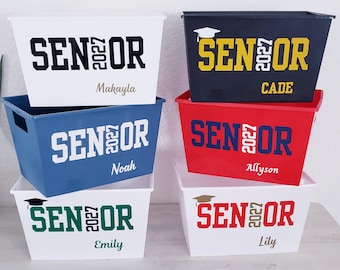 May include: Six rectangular plastic storage bins in white, black, blue, and red. Each bin features the text SENIOR with 2027 vertically between the letters. Names Makayla, Cade, Noah, Allyson, Emily, and Lily are printed on the respective bins.