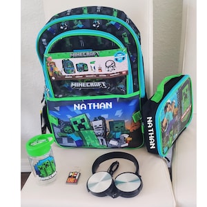 May include: A Minecraft-themed 5-piece backpack set featuring a black and green backpack with the name NATHAN, a matching lunch bag with the name NATHAN, a clear water bottle with a green lid, a small rectangular keychain, and black over-ear headphones.