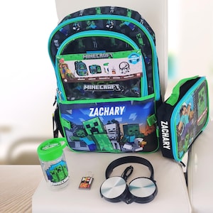 May include: A Minecraft 5-piece school set featuring a black and green backpack with the name ZACHARY, a matching lunch bag with the name ZACHARY, a clear water bottle with a Creeper graphic, a small rectangular keychain, and black headphones.
