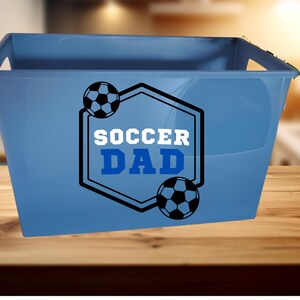 May include: A light blue plastic storage bin with integrated side handles. The front features a black hexagonal outline graphic containing two black and white soccer balls and the text "SOCCER DAD" in bold white and blue block lettering.