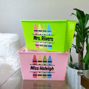 May include: Two plastic storage bins, one green and one pink, stacked on top of each other. Each bin has a crayon graphic and a name printed on the front. The green bin says "Mrs. Rivers" and the pink bin says "Miss Haleigh."