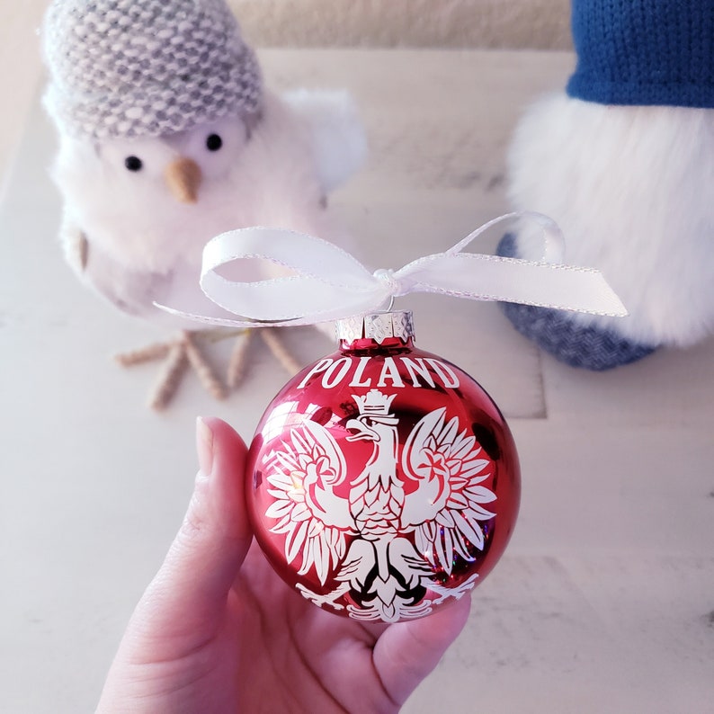 Op de afbeelding: Een rode kerstbal met het woord "POLAND" en een wit Pools adelaarsymbool. De ornament is rond, glanzend en heeft een wit lint om op te hangen. De achtergrond bevat een witte vogel met een grijze hoed.