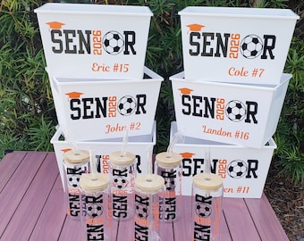 May include: A collection of white plastic storage bins and clear glass tumblers with bamboo lids and straws. Each item features black and orange text reading SENIOR 2026 with a soccer ball graphic, a graduation cap, and personalized names and jersey numbers.