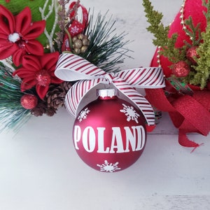 Op de afbeelding: Een rode kerstbal met het woord "POLAND" in het wit, versierd met sneeuwvlokken. De bal is vastgebonden met een gestreept rood-wit lint. Feestelijke vakantiedecoratie.