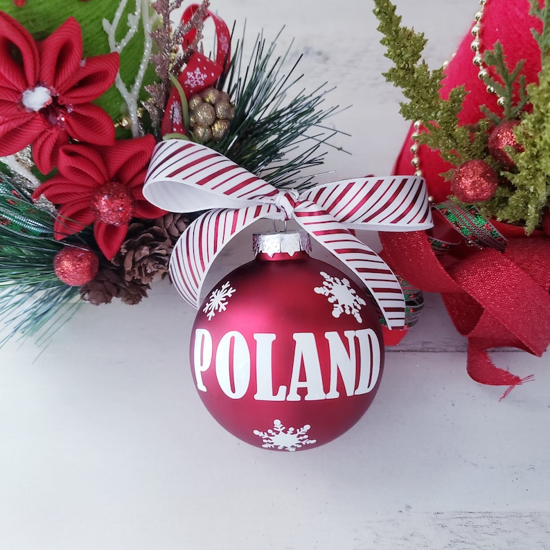 Op de afbeelding: Een rode kerstbal met het woord "POLAND" in het wit, versierd met sneeuwvlokken. De bal is vastgebonden met een gestreept rood-wit lint. Feestelijke vakantiedecoratie.
