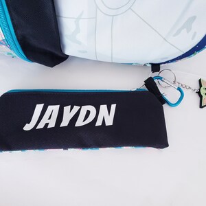 Pode incluir: Um estojo preto com o nome "JAYDN" em branco, um z&iacute;per azul e um padr&atilde;o colorido. Uma mochila com um design de Estrela da Morte e um chaveiro Baby Yoda tamb&eacute;m s&atilde;o vis&iacute;veis.
