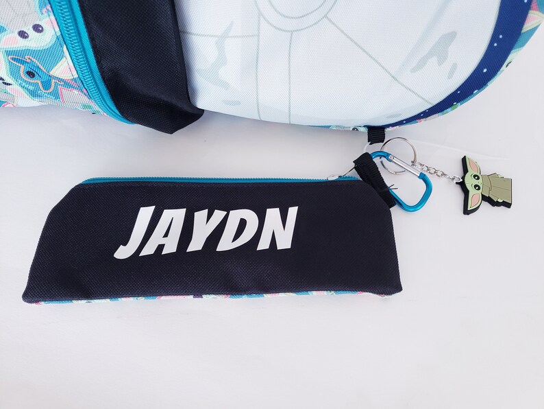 Pode incluir: Um estojo preto com o nome "JAYDN" em branco, um z&iacute;per azul e um padr&atilde;o colorido. Uma mochila com um design de Estrela da Morte e um chaveiro Baby Yoda tamb&eacute;m s&atilde;o vis&iacute;veis.