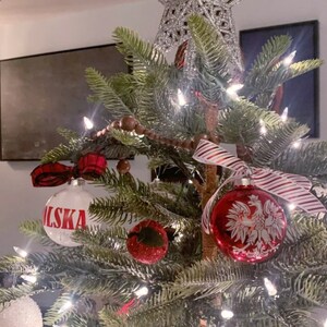 Op de afbeelding: Een versierde kerstboom met ornamenten, lichtjes en een ster. De ornamenten omvatten een witte bal met het woord "POLSKA" in het rood en een rode bal met een wit adelaarsontwerp. Er is ook een rood-wit gestreept lint aanwezig.