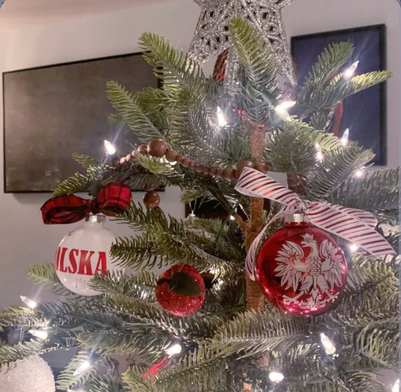 Op de afbeelding: Een versierde kerstboom met ornamenten, lichtjes en een ster. De ornamenten omvatten een witte bal met het woord "POLSKA" in het rood en een rode bal met een wit adelaarsontwerp. Er is ook een rood-wit gestreept lint aanwezig.
