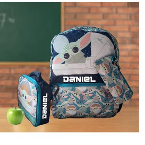 Pode incluir: Um conjunto de mochila, estojo e lancheira azul marinho com um design Grogu. A mochila e a lancheira t&ecirc;m o nome "DANIEL" em letras brancas. Uma ma&ccedil;&atilde; verde est&aacute; ao lado da lancheira.