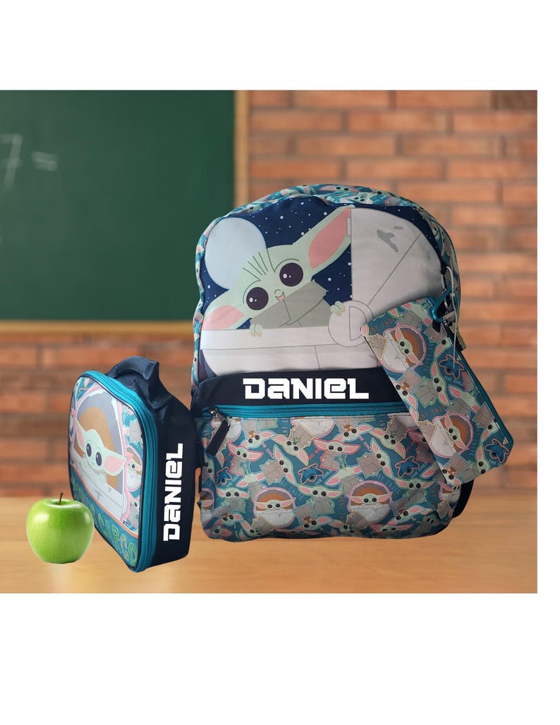 Pode incluir: Um conjunto de mochila, estojo e lancheira azul marinho com um design Grogu. A mochila e a lancheira t&ecirc;m o nome "DANIEL" em letras brancas. Uma ma&ccedil;&atilde; verde est&aacute; ao lado da lancheira.