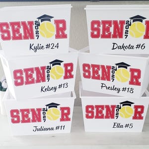 May include: White plastic storage bins with red "SENIOR 2027" text, a graduation cap, and a yellow softball. Each bin has a name and number: Kylie #24, Dakota #6, Kelsey #13, Presley #18, Juliana #11, and Ella #5.
