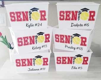 May include: White plastic storage bins with red "SENIOR 2027" text, a graduation cap, and a yellow softball. Each bin has a name and number: Kylie #24, Dakota #6, Kelsey #13, Presley #18, Juliana #11, and Ella #5.