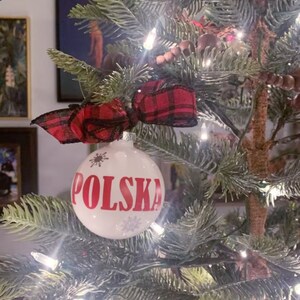 Op de afbeelding: Een witte kerstbal met het woord "POLSKA" in rode letters, versierd met sneeuwvlokken. De ornament is versierd met een rood-zwarte geruite strik en hangt aan een versierde kerstboom.