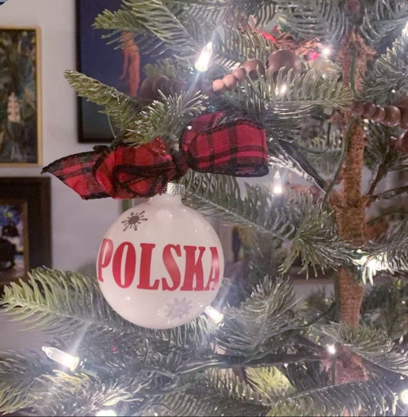 Op de afbeelding: Een witte kerstbal met het woord "POLSKA" in rode letters, versierd met sneeuwvlokken. De ornament is versierd met een rood-zwarte geruite strik en hangt aan een versierde kerstboom.