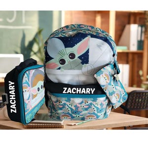 Pode incluir: Um conjunto de mochila de tr&ecirc;s pe&ccedil;as com design Grogu. O conjunto inclui uma mochila, uma lancheira e um estojo. A mochila e a lancheira t&ecirc;m o nome "ZACHARY" impresso. O conjunto &eacute; principalmente azul-petr&oacute;leo, azul e branco.