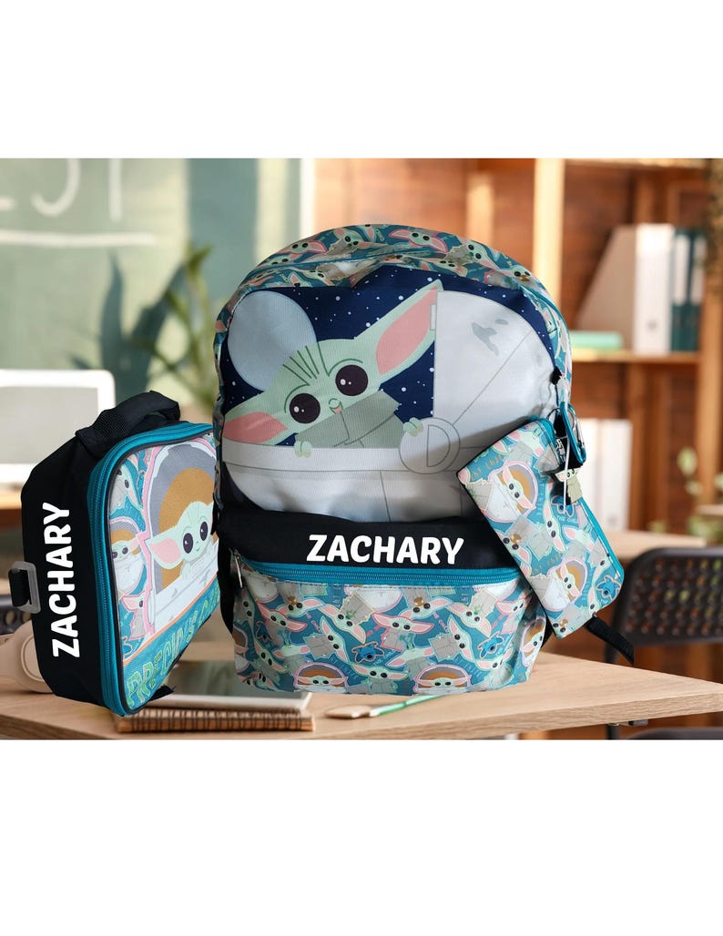 Pode incluir: Um conjunto de mochila de tr&ecirc;s pe&ccedil;as com design Grogu. O conjunto inclui uma mochila, uma lancheira e um estojo. A mochila e a lancheira t&ecirc;m o nome "ZACHARY" impresso. O conjunto &eacute; principalmente azul-petr&oacute;leo, azul e branco.