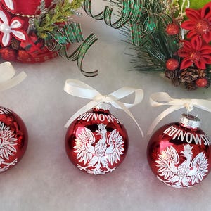 Op de afbeelding: Drie rode kerstballen met witte strikken. Elke bal heeft een wit Pools adelaarsymbool en het woord "BUINOWS" in het wit. Feestelijke vakantiedecoratie.