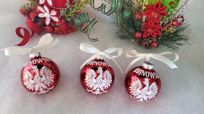 Op de afbeelding: Drie rode kerstballen met witte strikken. Elke bal heeft een wit Pools adelaarsymbool en het woord "BUINOWS" in het wit. Feestelijke vakantiedecoratie.