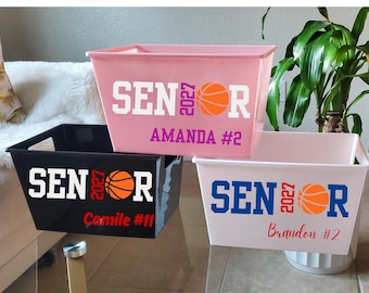 Puede incluir: Tres cajas de almacenamiento rectangulares de plástico en rosa, negro y blanco. Cada caja presenta el texto SENIOR 2027 con un gráfico de baloncesto naranja que reemplaza la letra O, y un nombre y número personalizados en color.