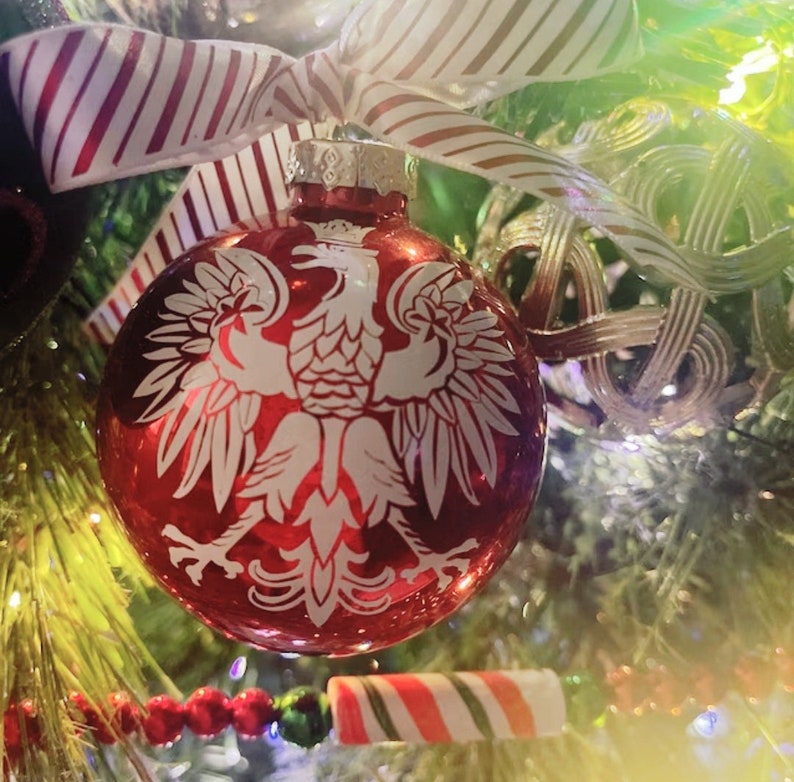 Op de afbeelding: Een rode glazen kerstbal met een wit Pools adelaarsymbool. De ornament hangt aan een versierde kerstboom met een gestreept lint en zuurstokversieringen.